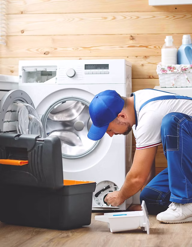 A repairman in blue overalls and a cap kneels in front of a washing machine, using a tool to fix it. A toolbox is open nearby, and detergent bottles sit on a shelf—professional Appliance Repair in Oakland CA in action.