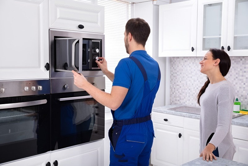 A repairman in blue overalls provides appliance repair in Oakland CA, fixing a microwave in a modern white kitchen while a woman stands nearby, smiling and watching him work.
