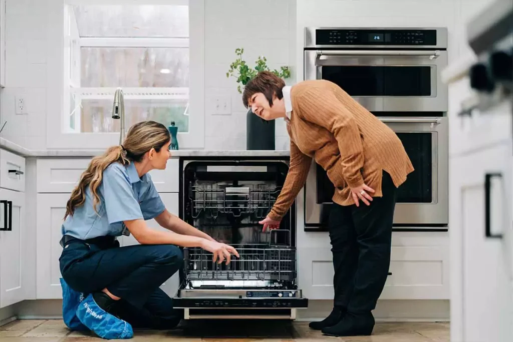 Two women in a kitchen examine the inside of an open dishwasher, possibly discussing Appliance Repair in Oakland CA. One kneels in work clothes while the other stands, pointing at the racks. White cabinets and built-in ovens are visible behind them.
