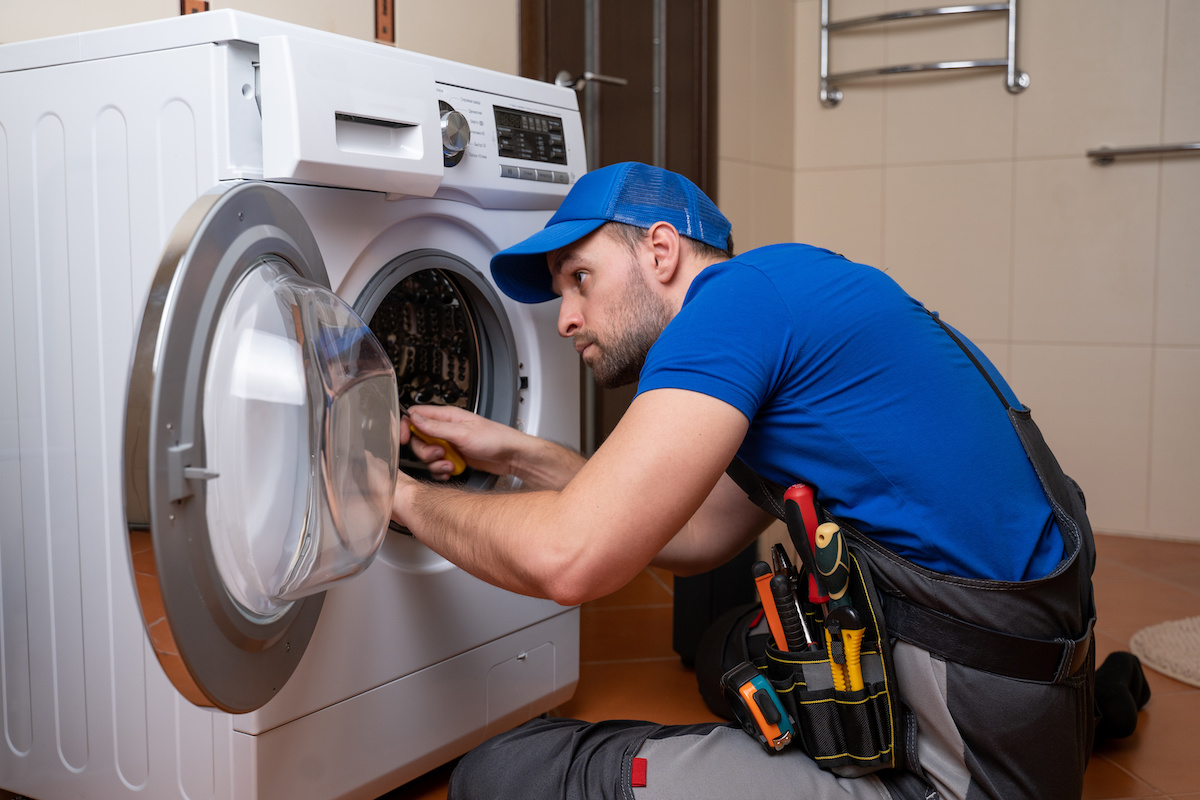 A repairman in a blue cap and shirt is kneeling next to a front-loading washing machine, performing Appliance Repair in Oakland CA. He uses a screwdriver and tool belt while working in a tiled laundry room.