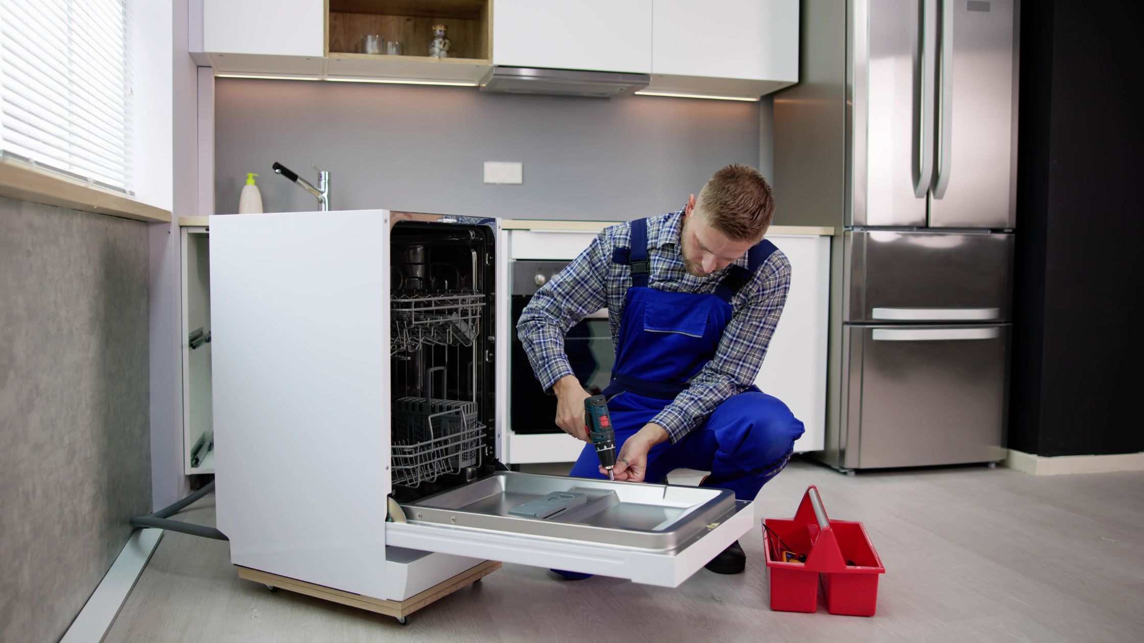 A repairman in blue overalls kneels on a kitchen floor, using a screwdriver to fix an open dishwasher. A red toolbox sits nearby, showcasing expert Appliance Repair in Oakland CA among modern appliances and light-colored cabinets.