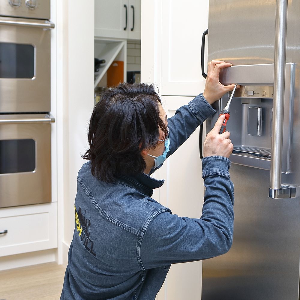 A person wearing a mask and blue shirt uses a screwdriver to repair the ice and water dispenser on a stainless steel refrigerator in a modern kitchen, showcasing expert Appliance Repair in Oakland CA.